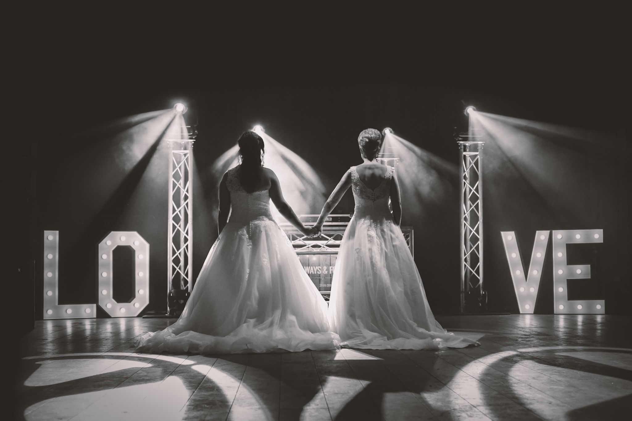 Two brides holding hands before an illuminated LOVE sign